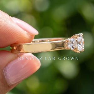 May include: A gold ring featuring a round brilliant-cut centre stone and two smaller side stones. The band is smooth and polished. The text BENIYAZ LAB GROWN is printed in white on the lower portion of the image, centred horizontally.