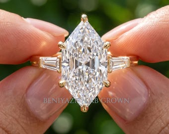 May include: A gold ring featuring a large marquise-cut center stone held by six prongs, flanked by two tapered baguette-cut side stones. The text BENIYAZI LAB GROWN is visible on the lower portion of the center stone setting in this jewelry piece.