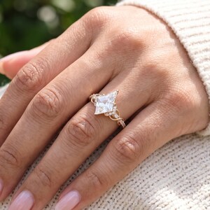 A woman's hand wearing a geometric kite cut diamond engagement ring, resting on a soft cream knitted fabric. The prominent kite shaped center diamond and sparkling pavé band create an elegant bridal look, with natural skin texture and neatly manicured nails.