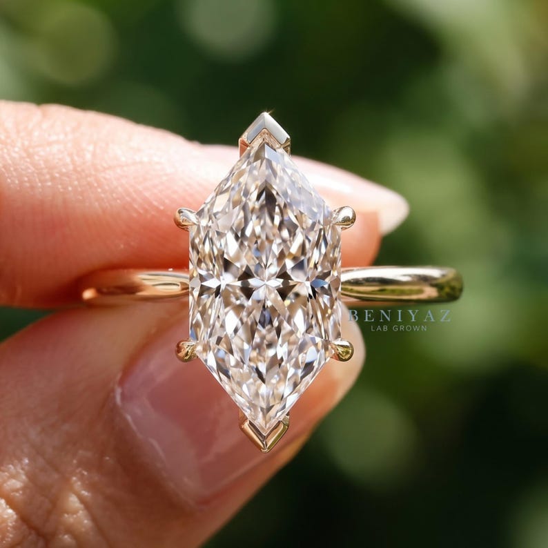 Op de afbeelding: Een ring met een diamant in marquise-slijpvorm, gezet in een gepolijste geelgouden band. De centrale steen heeft een gefacetteerde, langwerpige vorm en wordt vastgehouden door zes pootjes. De tekst BENIYAZ LAB GROWN is zichtbaar op de zijkant.