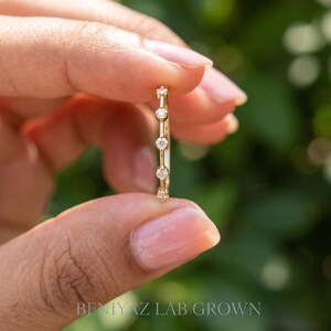 Delicate marquise moissanite stacking ring held vertically between fingers. The side angle shows the raised gemstone settings and smooth polished band against a blurred green garden background.