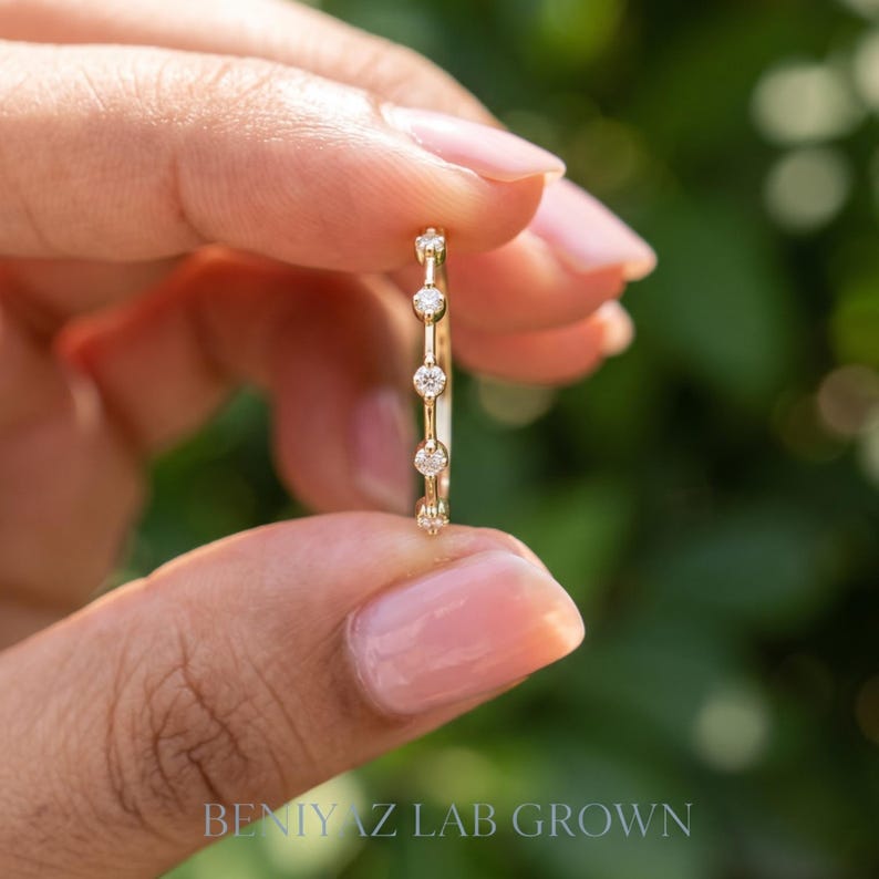 Delicate marquise moissanite stacking ring held vertically between fingers. The side angle shows the raised gemstone settings and smooth polished band against a blurred green garden background.