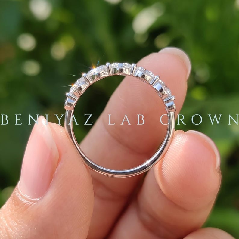 Rear view of a marquise lab grown diamond band held between fingertips. The photograph shows the smooth rounded white metal shank, slim band construction and elevated marquise diamond setting against a blurred green garden background.