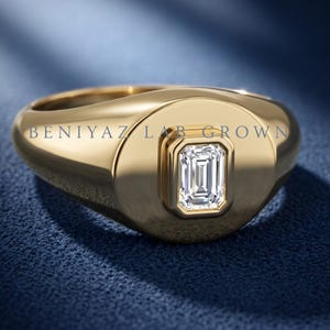 Yellow gold emerald cut lab grown diamond signet ring on blue fabric, showing the rounded face and tapered shoulders.