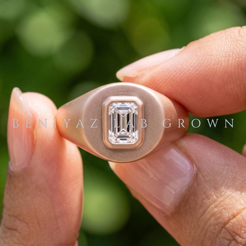 Hand held close-up of a rose gold emerald cut diamond signet ring showing the rounded face and center stone.