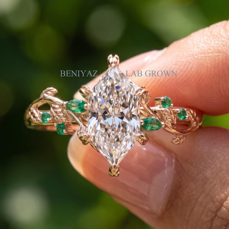 Close up hand held view of a Dutch marquise lab grown diamond engagement ring with green accent stones and detailed vine and leaf shoulders.