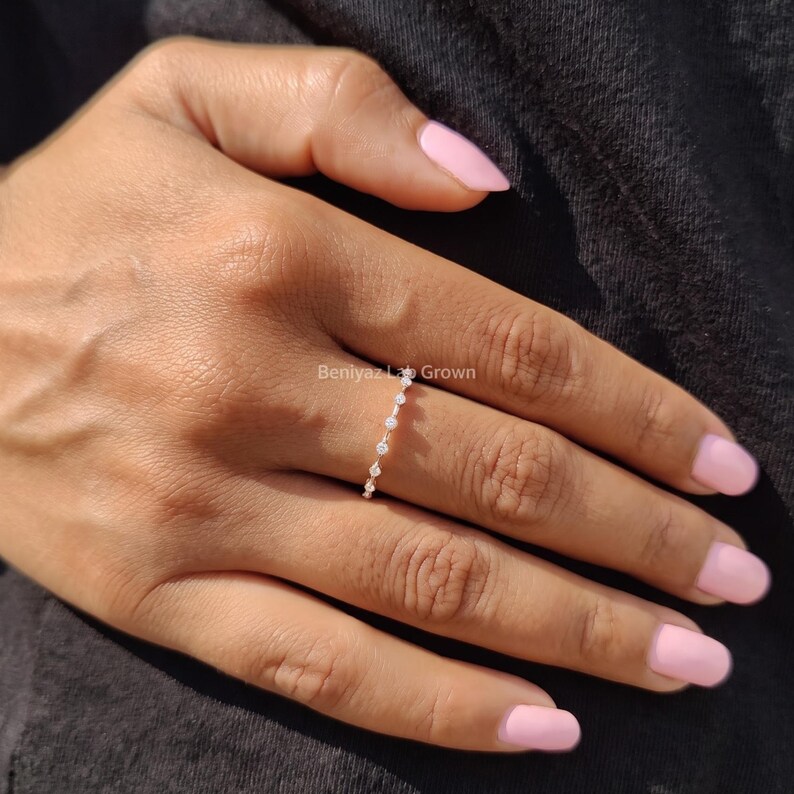 Woman wearing a delicate marquise moissanite wedding band on her finger. The slim ring features sparkling stones and is styled with natural pink nails and a dark textured sleeve.