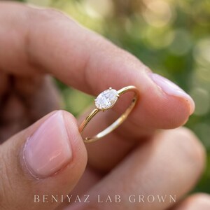 Rear angled view of a minimalist oval diamond ring held between manicured fingertips. The smooth yellow gold shank and delicate bezel setting are visible, showing the simple construction and slender proportions of the ring.