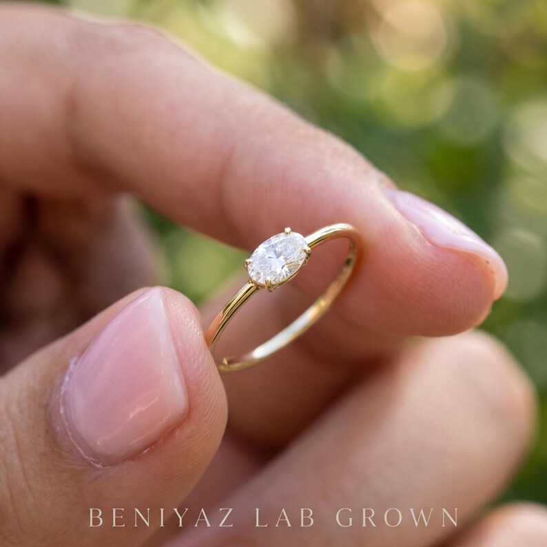 Rear angled view of a minimalist oval diamond ring held between manicured fingertips. The smooth yellow gold shank and delicate bezel setting are visible, showing the simple construction and slender proportions of the ring.