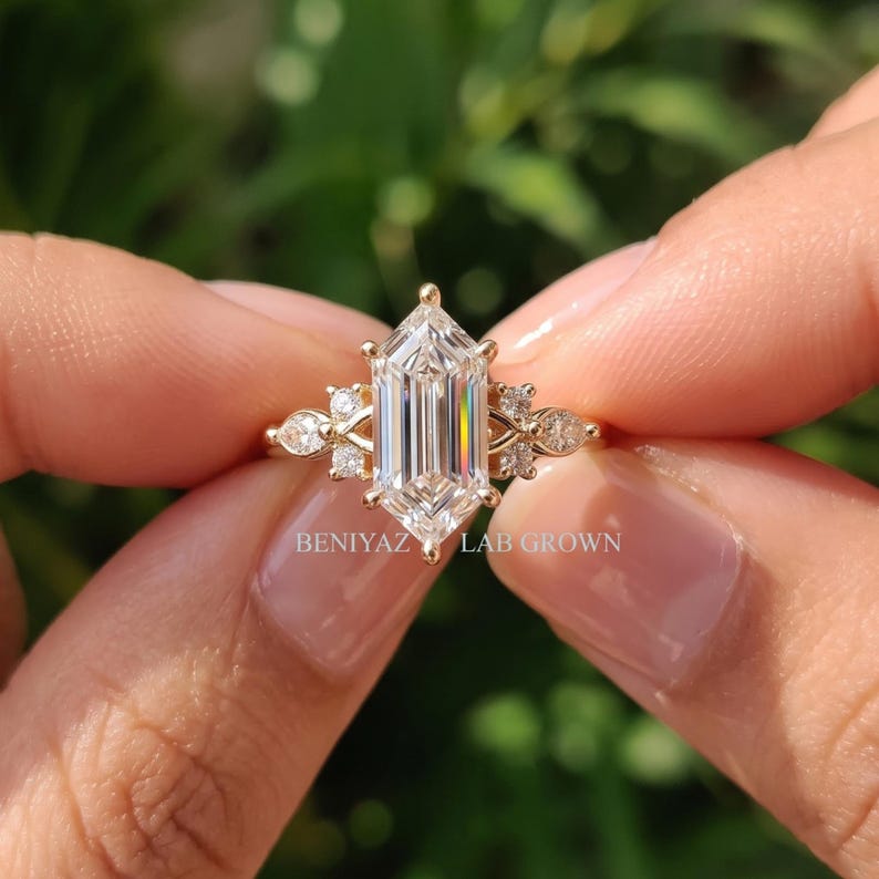 Close up geometric hexagon step cut engagement ring held by hand with lab grown diamond center and side stone cluster