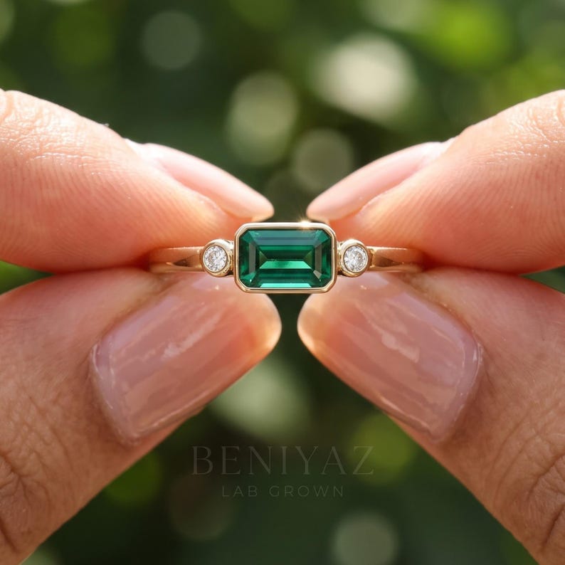 Front close up of three stone emerald engagement ring with east west green center stone and two lab grown diamonds in solid gold.