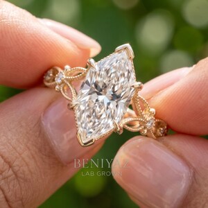 May include: A gold ring featuring a large marquise-cut center stone with a faceted surface. The band displays intricate filigree metalwork with small round accent stones embedded along the sides. The text BENIYAZ LAB GROWN is visible.