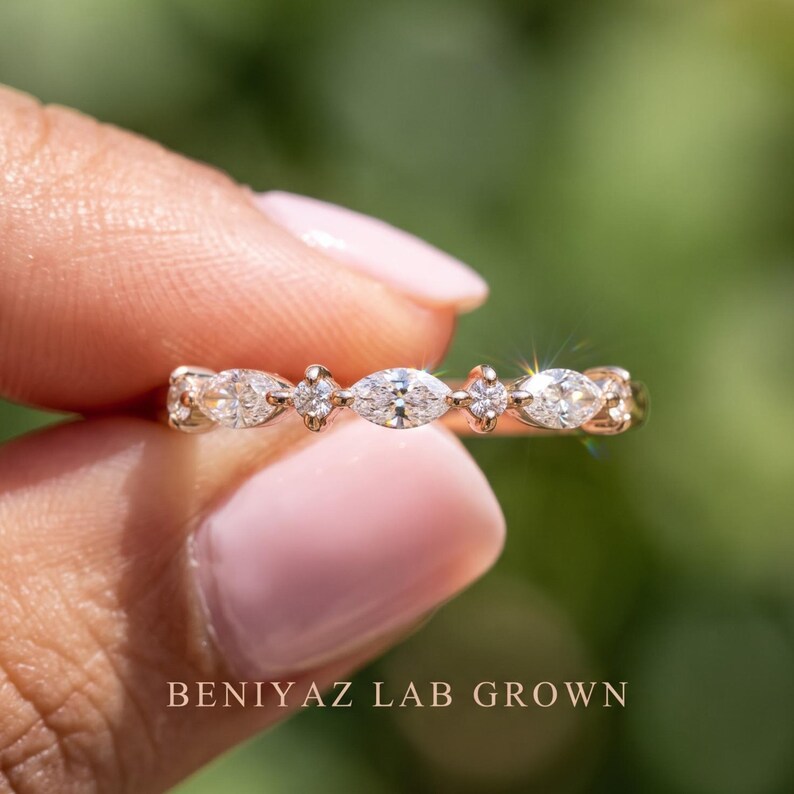 Macro close-up of a dainty marquise and round lab grown diamond ring held between fingertips. Alternating horizontally positioned marquise shaped diamonds and smaller round accents form a delicate sparkling pattern along the upper band. The softly blurred green background emphasizes the white gemstones, prong details and polished metal finish.