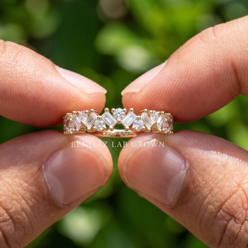 Hand holding a gold half eternity wedding band with baguette lab grown diamonds and cluster accents, modern stacking ring photographed against green foliage
