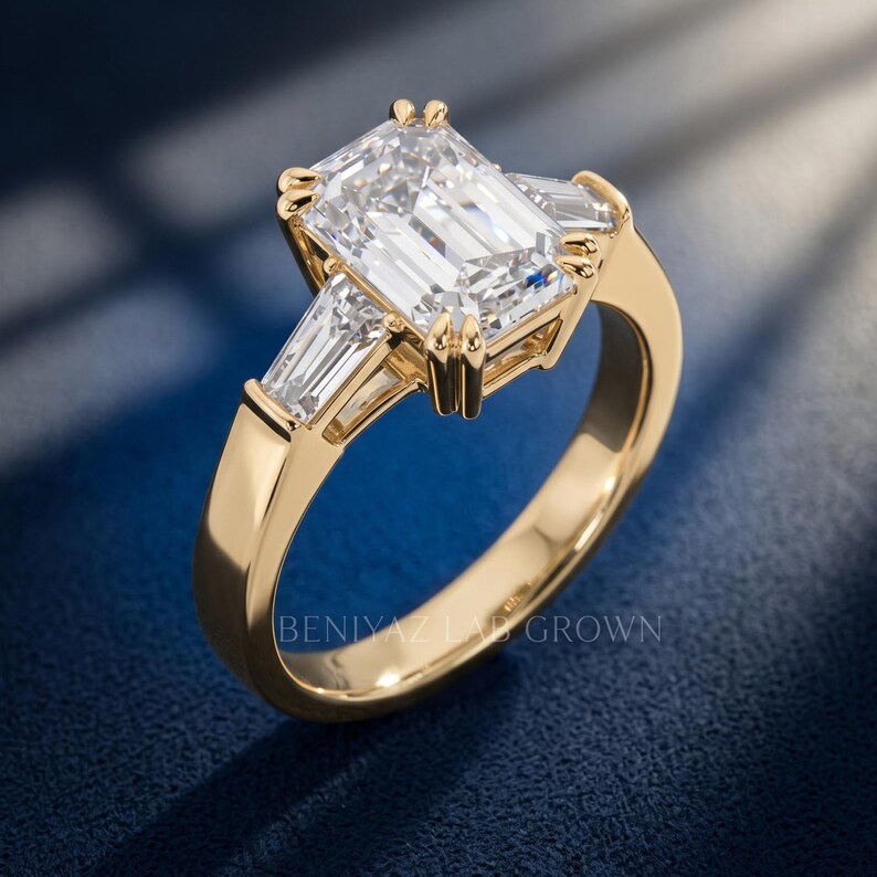 Angled view of emerald cut lab grown diamond engagement ring with tapered side stones, prong setting and polished yellow gold band