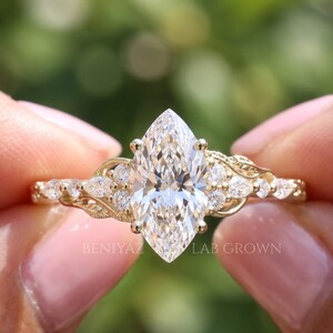 Detailed front view of a marquise lab grown diamond engagement ring held between fingertips. The brilliant center diamond is framed by smaller leaf inspired diamond accents extending along the slender yellow gold band, creating an elegant nature inspired bridal design.