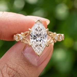 May include: A gold ring featuring a large marquise-cut center stone held by a prong setting. The band displays intricate filigree metalwork and is accented with small round stones embedded along the sides of the shank in a symmetrical pattern.