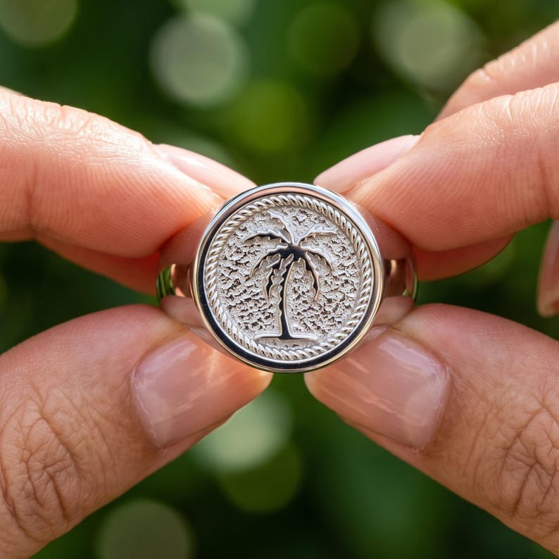 Close up hand photo of a sterling silver palm tree signet ring with textured round face, raised tropical palm motif and rope-style border