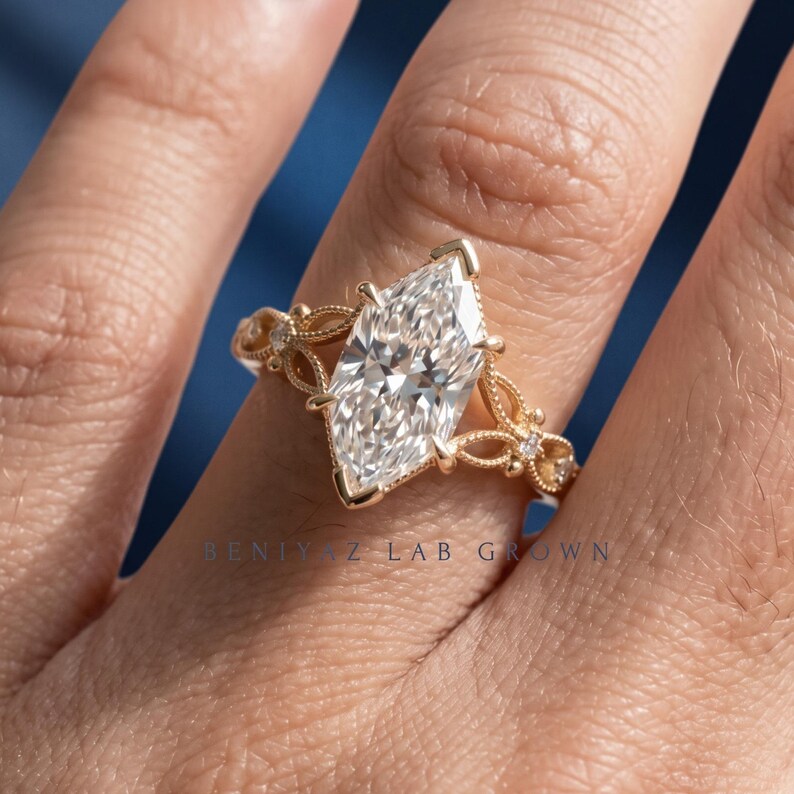 May include: A gold ring featuring a large marquise-cut center stone held by prongs. The band displays intricate filigree metalwork with small accent stones embedded along the sides. The text BENIYAZ LAB GROWN is visible on the ring band.