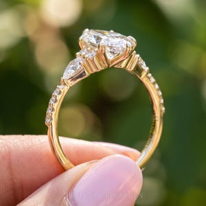 Full circular profile of a marquise engagement ring held between fingertips outdoors. The photograph shows the raised diamond cluster, decorative gallery, accent stones along the shoulders and polished gold tone band.