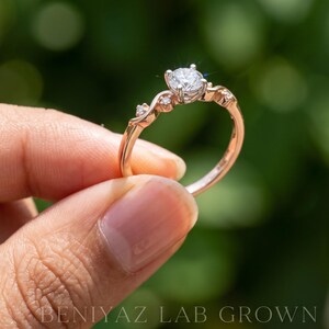 K&ouml;nnte beinhalten: Ein Ring aus Ros&eacute;gold mit einem zentralen, rund geschliffenen wei&szlig;en Stein in einer Vier-Krappen-Fassung, flankiert von zwei kleineren runden Steinen auf jeder Seite der Ringschiene. Der Text BENIYAZ LAB GROWN ist unten im Bild zu sehen.