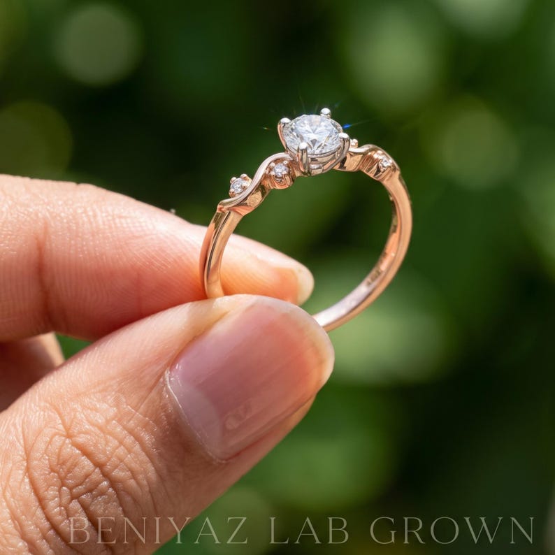 K&ouml;nnte beinhalten: Ein Ring aus Ros&eacute;gold mit einem zentralen, rund geschliffenen wei&szlig;en Stein in einer Vier-Krappen-Fassung, flankiert von zwei kleineren runden Steinen auf jeder Seite der Ringschiene. Der Text BENIYAZ LAB GROWN ist unten im Bild zu sehen.