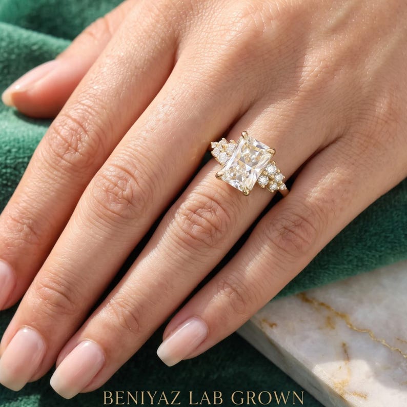 May include: A gold ring featuring a large rectangular radiant-cut center stone flanked by a cluster of three smaller round-cut stones on each side. The band is thin and polished gold. The text BENIYAZ LAB GROWN is printed at the bottom of the image.