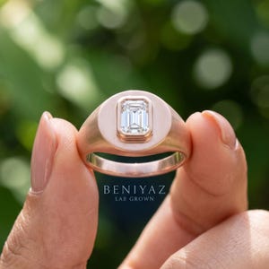 Silver tone emerald cut lab grown diamond signet ring held between fingers with a smooth polished statement design.