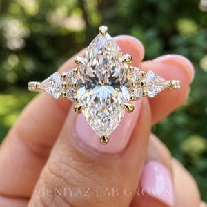 Detailed frontal photograph of a marquise lab grown diamond engagement ring supported by fingertips. The sparkling pointed center stone, geometric side diamonds and delicate gold tone setting stand out against blurred greenery.