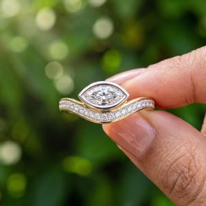 Close up hand view of a marquise lab grown diamond ring in yellow gold featuring a vintage inspired halo and pavé diamond band against soft green foliage