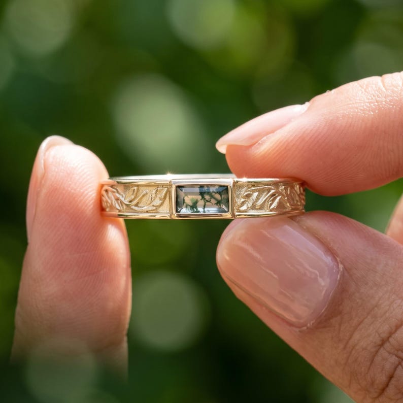 Puede incluir: Un anillo de oro con una piedra preciosa rectangular de &aacute;gata musgosa engastada en el centro. La banda presenta un patr&oacute;n en relieve detallado de hojas y enredaderas que se extiende desde el engaste por toda la superficie del metal.