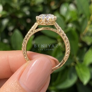 Three-quarter side view of an oval diamond engagement ring held between fingertips. The raised gemstone setting and delicate diamond detailing are visible along the yellow gold tone band.