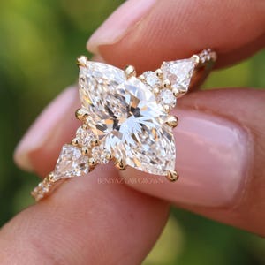 Close-up of a vintage inspired marquise engagement ring held between fingertips. The brilliant pointed center diamond is framed by kite shaped side stones and smaller accents, creating a symmetrical cluster design.