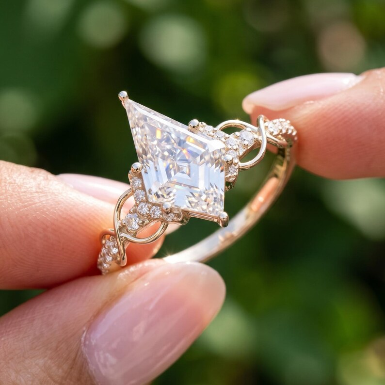 Angled macro photograph of a kite cut diamond engagement ring held between fingertips outdoors. The side perspective reveals the pointed diamond outline, raised center setting, curved accent stones and intricate gold tone infinity inspired band against green foliage.