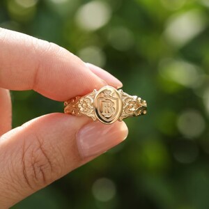 May include: A gold ring featuring an oval-shaped face with an engraved shield design containing the letters B and B. The band is decorated with intricate floral carvings on both sides of the central oval emblem, showcasing a polished finish.