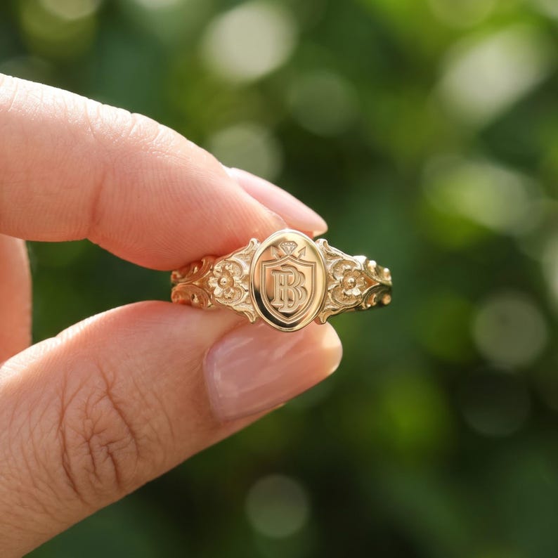 May include: A gold ring featuring an oval-shaped face with an engraved shield design containing the letters B and B. The band is decorated with intricate floral carvings on both sides of the central oval emblem, showcasing a polished finish.