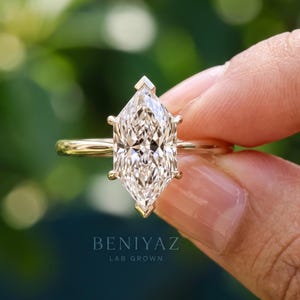 Op de afbeelding: Een gouden ring met een centraal geslepen marquise steen, vastgehouden door zes pootjes. De ringband is van glad en gepolijst goud. De tekst BENIYAZ LAB GROWN is onderaan in beeld zichtbaar. Het ontwerp richt zich op de langwerpige steen.