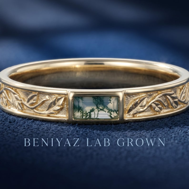 Puede incluir: Un anillo de oro con una piedra de &aacute;gata musgosa rectangular con inclusiones verdes, engastada entre bandas grabadas con un patr&oacute;n de hojas. El texto BENIYAZ LAB GROWN es visible debajo del anillo, que tiene un acabado pulido.