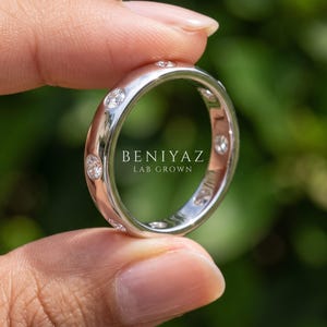 Close up hand view of a white gold lab grown diamond band with round bezel set diamonds, contemporary unisex wedding ring with green outdoor background