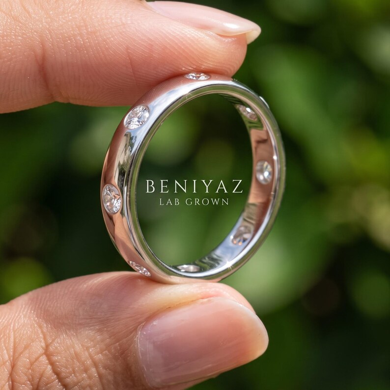 Close up hand view of a white gold lab grown diamond band with round bezel set diamonds, contemporary unisex wedding ring with green outdoor background