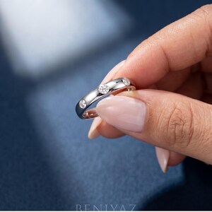 May include: A polished silver-toned band ring featuring three round, clear, bezel-set stones embedded along the top surface. The metal has a smooth, reflective finish, and the stones are spaced evenly across the curve of the circular band.