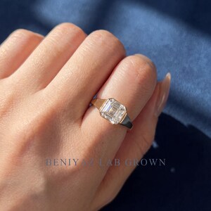 Angled close-up of a female hand wearing a 1.55 CT Asscher cut lab grown diamond engagement ring in 14K yellow gold. The bezel set signet design highlights an IGI certified D color VVS1 lab grown diamond, with the polished yellow gold band creating a bold and elegant fine jewelry style.