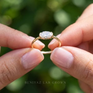 Side-angle photograph of a dainty oval lab grown diamond bezel ring held between two fingertips. The east west center stone sits across the thin polished gold band, showing the setting height and delicate proportions.