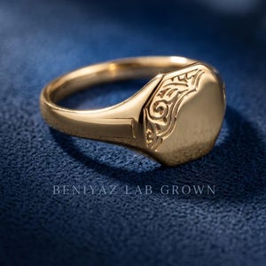 Yellow gold oval signet ring on blue background with polished engraving face, raised floral scrollwork and vintage inspired design