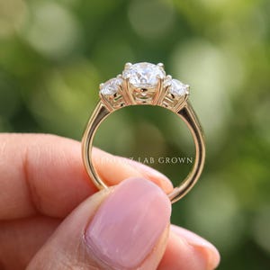 May include: A gold ring featuring a large central round-cut stone flanked by two smaller round-cut stones on each side. The band is smooth and polished gold. The text BENIYAZ LAB GROWN is visible on the inner surface of the ring band.
