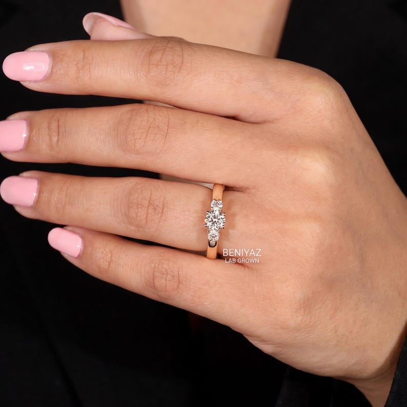 May include: A rose gold ring featuring three round-cut white stones set in a row. The centre stone is larger than the two flanking stones. The text BENIYAZ LAB GROWN is printed in white on the band near the setting of the ring.