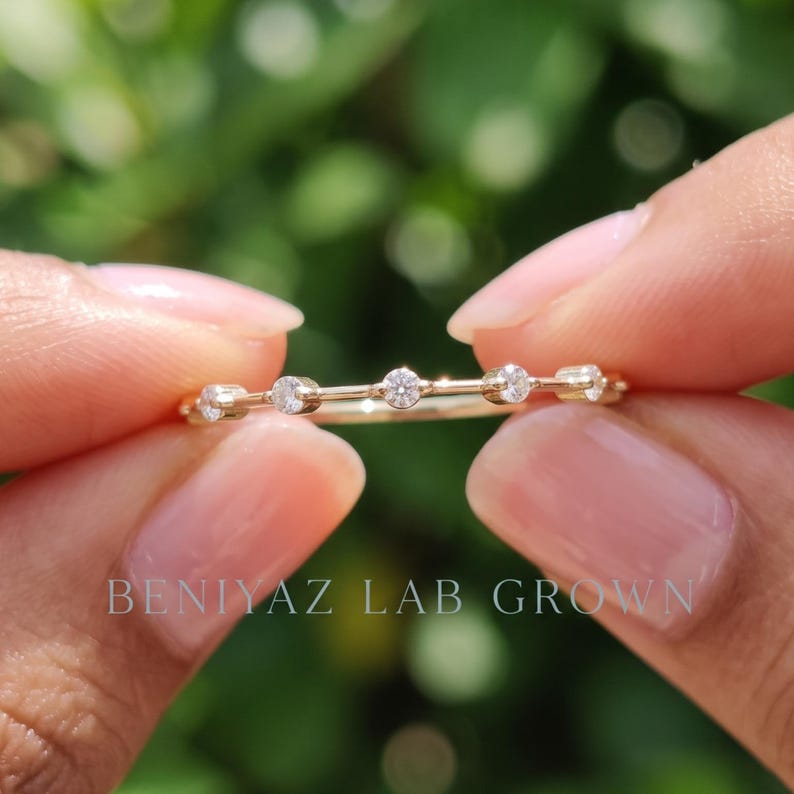 Angled close-up of a marquise moissanite stacking ring held between fingers. The photograph shows the slim band, sparkling stones and delicate setting against a natural green bokeh background.