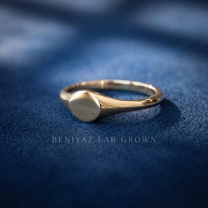 Yellow gold tone round signet ring on blue background with small plain face, minimalist tapered shank and polished finish