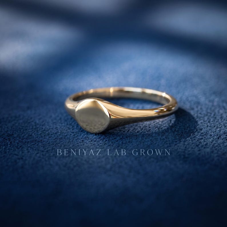 Yellow gold tone round signet ring on blue background with small plain face, minimalist tapered shank and polished finish