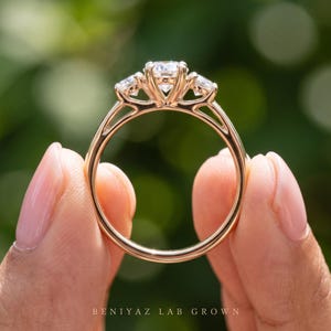 May include: A three-stone ring crafted from rose gold, featuring a large central round-cut white gemstone flanked by two smaller round-cut white gemstones. The band is smooth and polished, with the text BENIYAZ LAB GROWN printed at the base.
